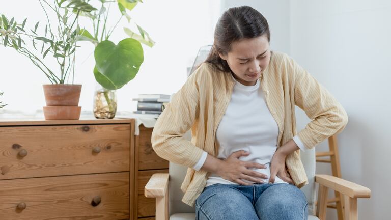 Una mujer sentada tocándose el abdomen con expresiones de dolor intenso.